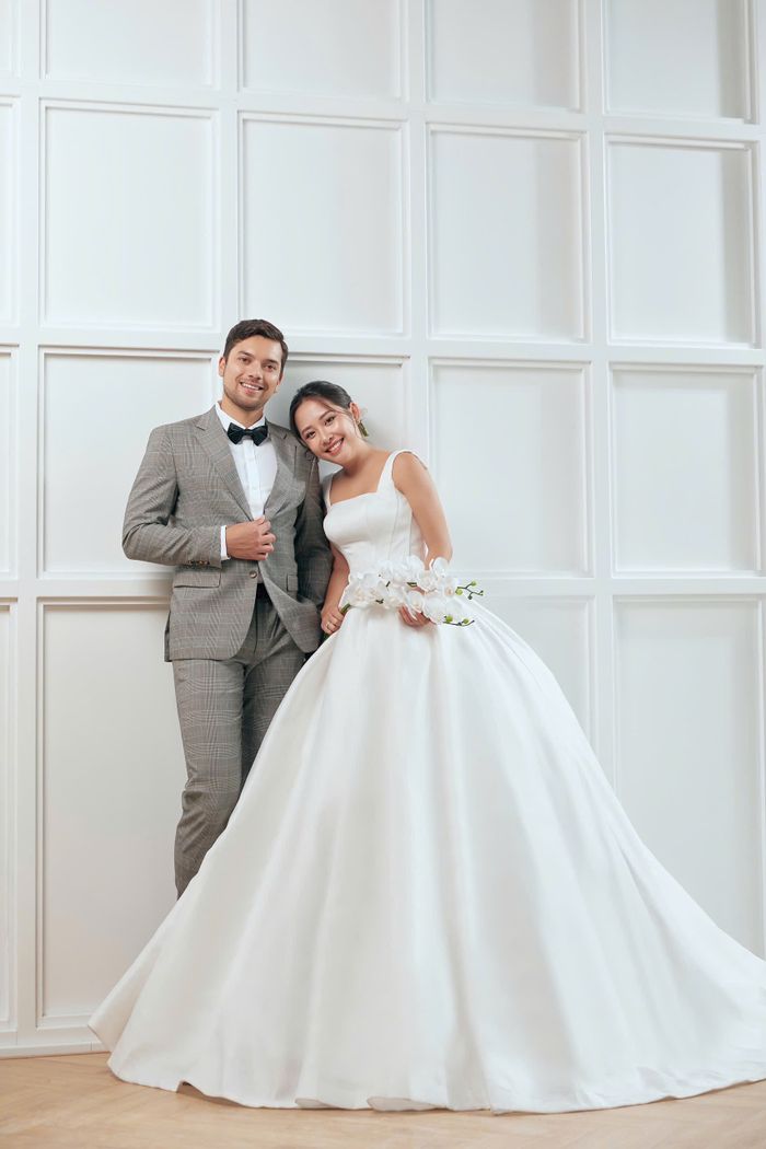 Smiling bride and groom in elegant attire posing indoors against a white paneled wall.